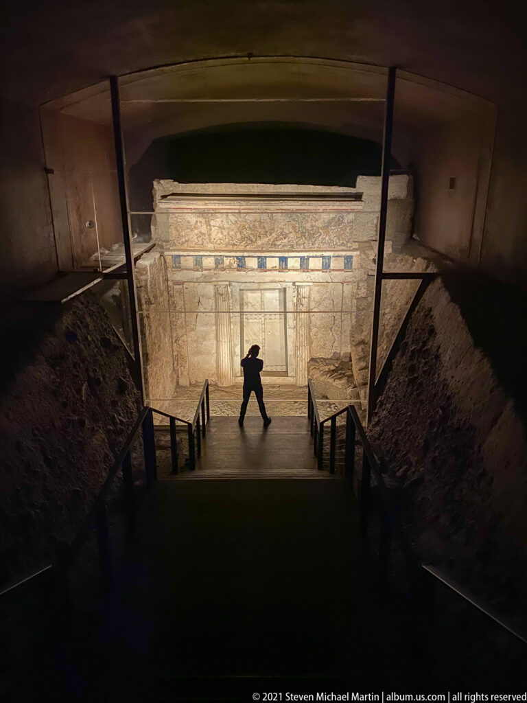 Wide view down a dark museum stair toward a lit Macedonian tomb façade, with a visitor silhouetted on the landing.