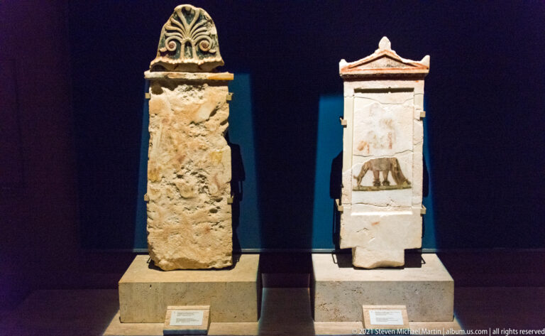 Two upright stone grave markers, one topped by a carved palmette and one by a small pediment with faded painted decoration.