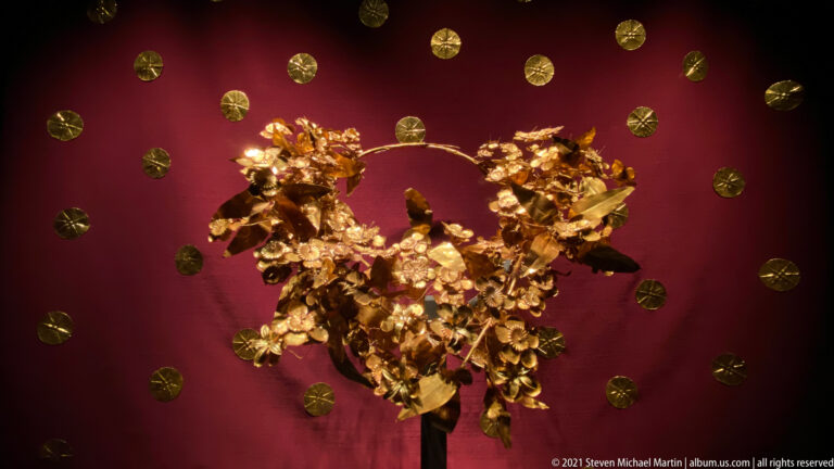 Gold oak wreath from the Tomb of Philip II mounted against red fabric with delicate leaves, acorns, flowers, and branching stems.
