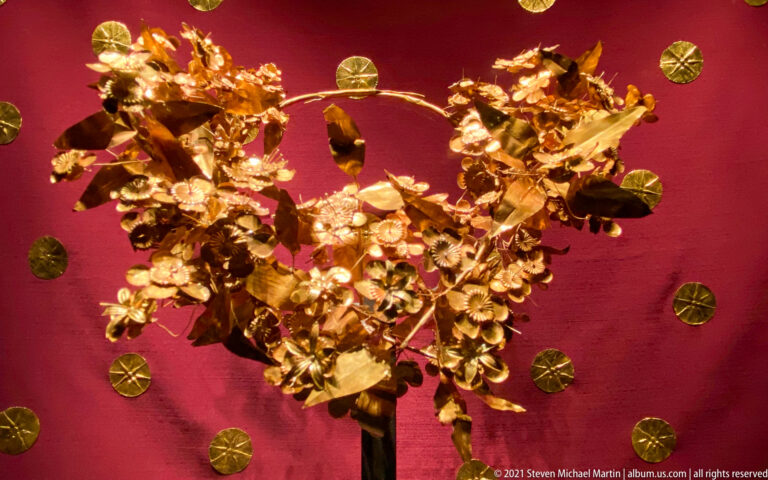 Close frontal view of a dense gold leaf wreath against red fabric, with circular gold appliqués visible around its edges.