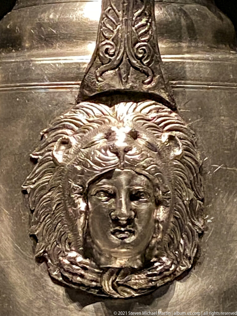 Silver vessel detail with a frontal relief mask framed by radiating forms beneath an ornamented vertical handle strap.