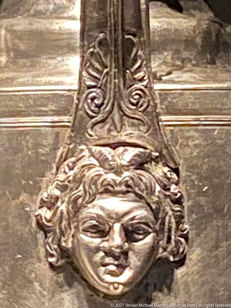 Close-up of a silver vessel handle attachment shaped as a human mask with curling ornament and engraved strap above.