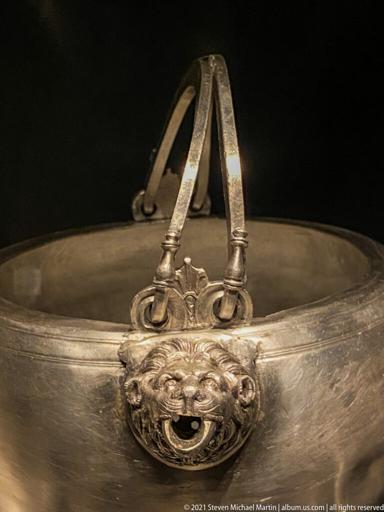 Large silver basin with an arched handle attached above a projecting animal-mask ornament on the rim.