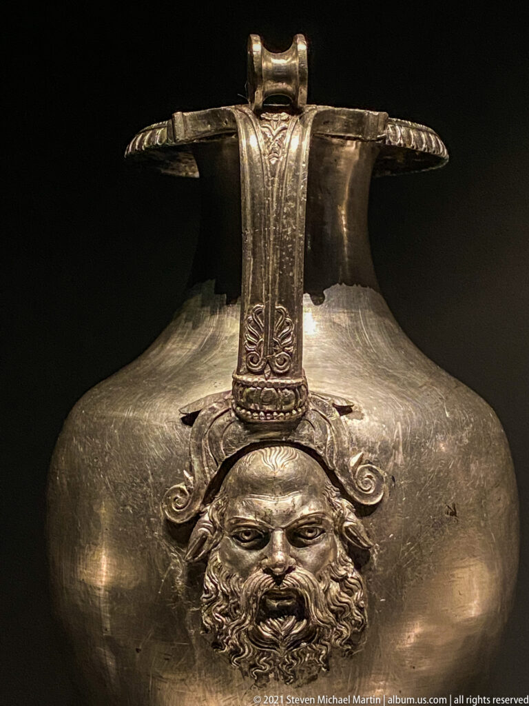 Large silver vessel with a bearded relief mask beneath a broad engraved handle strap across its rounded body.
