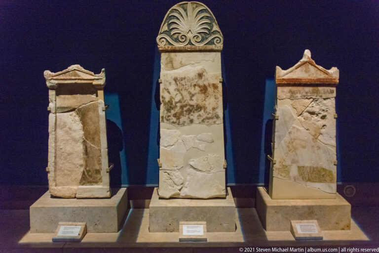 Three upright stone grave markers, including a tall central stele crowned by a large carved palmette.