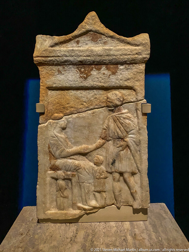 Rectangular stone relief with two facing draped figures beneath a molded upper frame and smaller carved forms below.