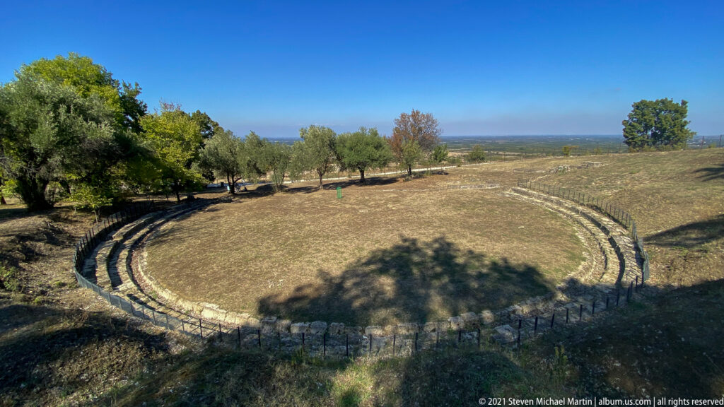Wide elevated view of the ancient theater at Aigai with semicircular stone seating, grassy center, trees, and distant plain.