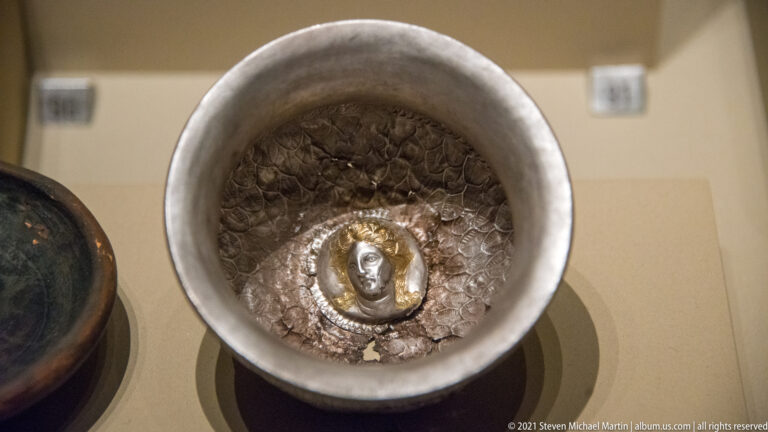 Top view into a silver-colored bowl with textured interior and a small gilded human-head medallion at the center.