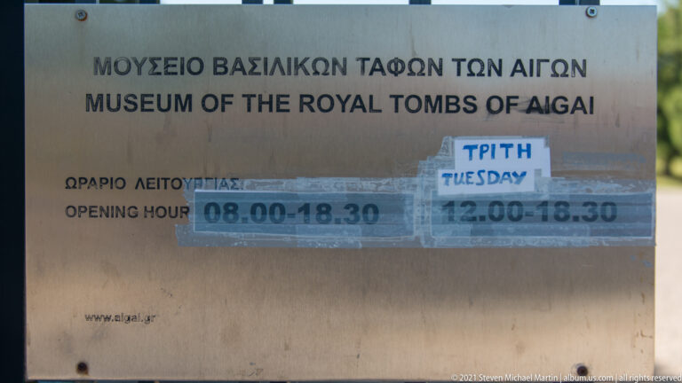 Bilingual Museum of the Royal Tombs of Aigai sign with posted opening hours and temporary paper notices.