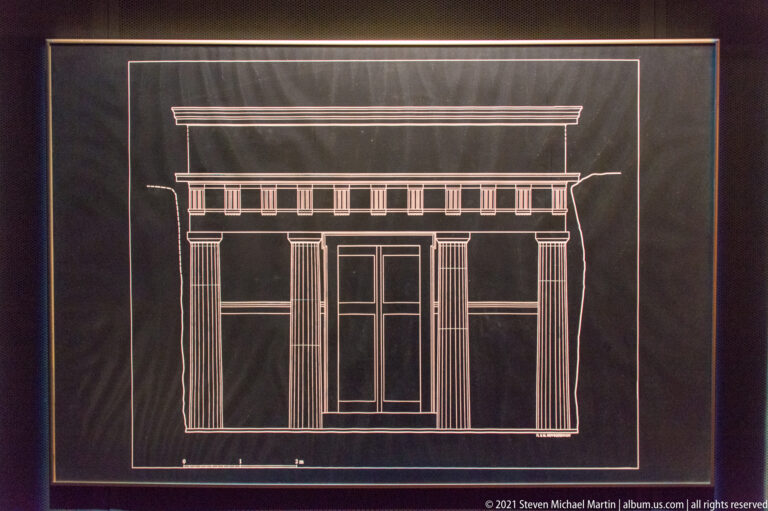 White line elevation on a dark panel showing a symmetrical tomb façade with central double door and flanking supports.
