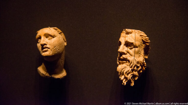 Two small carved male heads mounted on a dark wall, one youthful and one bearded with deeply modeled hair.