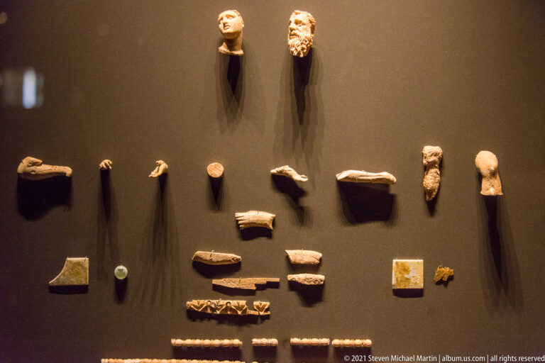 Museum wall display of two carved male heads and many small fragmentary hands, limbs, plaques, and decorative pieces.
