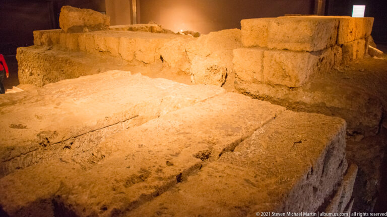 Large pale stone slabs and low block walls forming excavated foundations under warm indoor lighting.