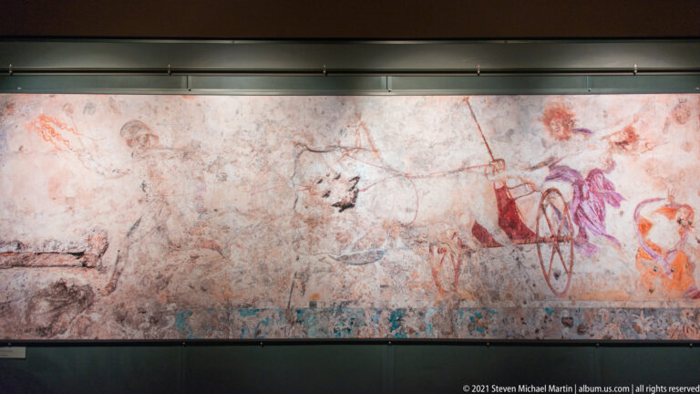 Wide ancient wall painting with faded red and violet figures spread across worn plaster, with denser figural action at right.