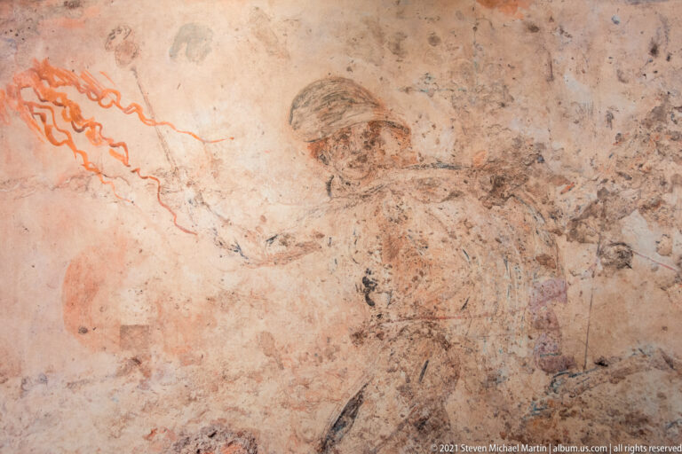 Faded standing male figure outlined in brown on worn plaster, with red linear decoration surviving along the left edge.