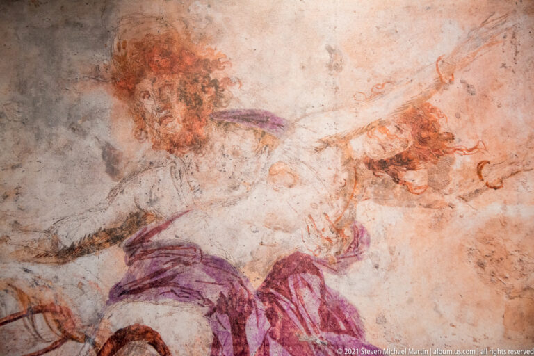 Close view of a worn ancient hunt painting with violet-clad riders, horses, and reddish animals overlapping across pale plaster.