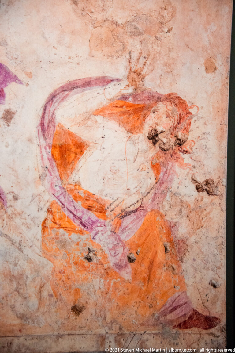 Mounted hunter in orange with a sweeping violet cloak rides across a faded ancient wall painting.