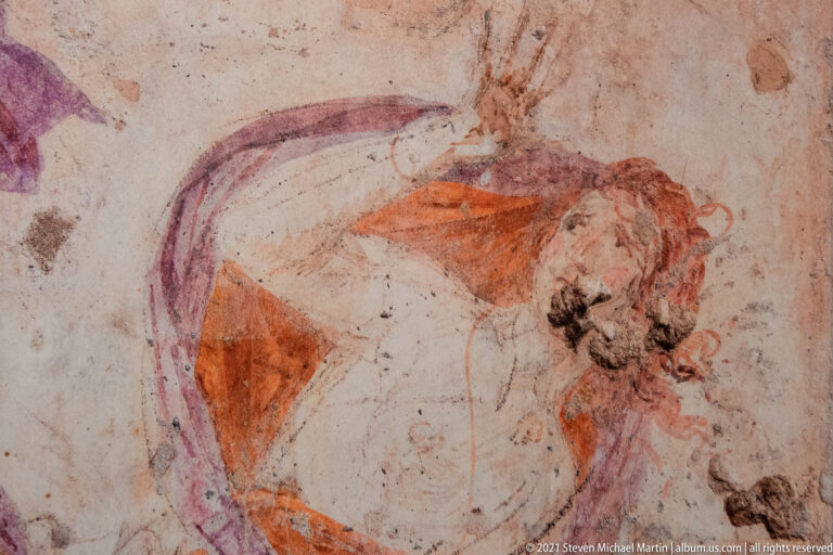Close detail of a mounted figure leaning forward, with orange clothing and violet drapery on worn pale plaster.