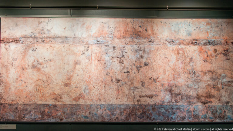Long, heavily worn painted wall with faint reddish figures and a darker horizontal decorative band along the bottom.