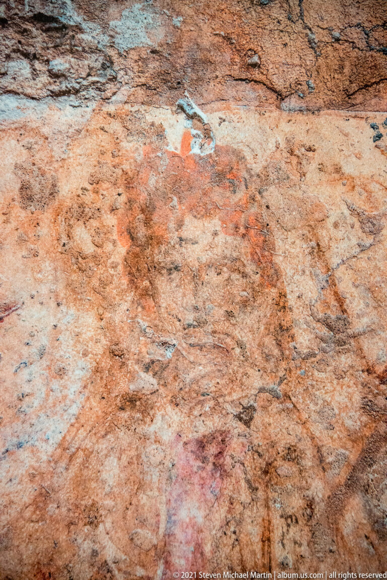Close view of rough ancient plaster with scattered red-orange and blue-green pigment and faint curving outlines.
