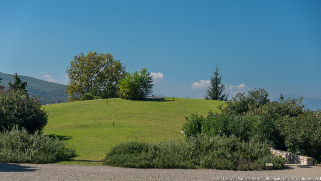 Grass-covered Great Tumulus rising above shrubs and trees under a clear blue sky at Aigai.