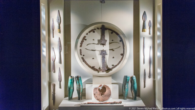 Museum case with a large circular object, central weapon, spearheads, turquoise-patinated vessels, and a ceramic container.