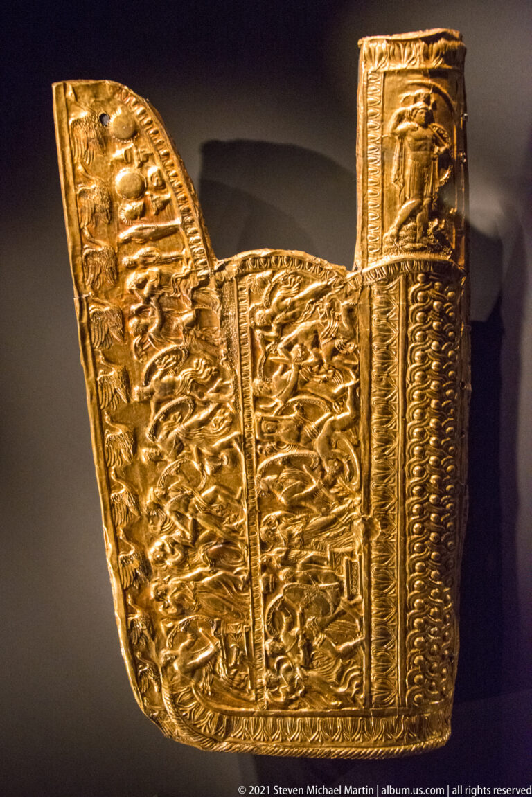 Curved gold sheet fitting covered with embossed figures, animals, borders, and dense ornamental relief.