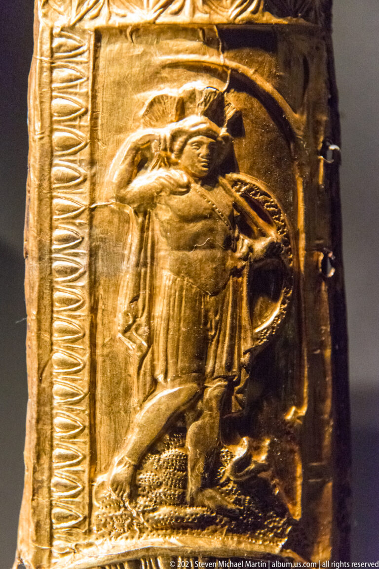 Close view of a small gold armor fitting with a standing figure modeled in repoussé inside an ornamental border.