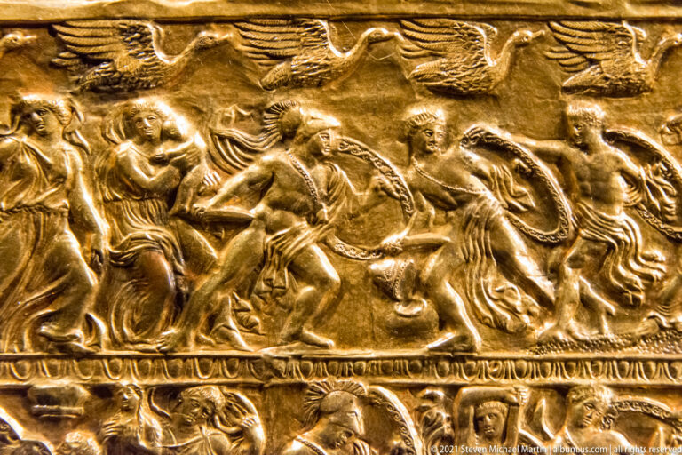Embossed gold fitting with a crowded relief scene of seated, standing, and winged figures.