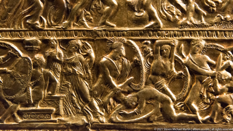 Close-up of an embossed gold relief with several overlapping human figures beneath a narrow repeating ornamental border.