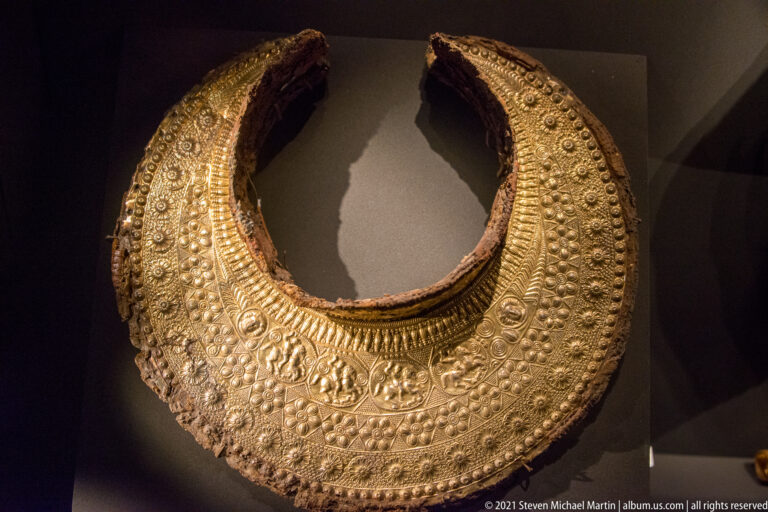 Large crescent-shaped gold ornament densely decorated with rosettes, bosses, beading, and a band of figural medallions.