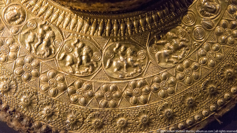 Close detail of gold repoussé work with rosettes, spirals, beaded borders, and circular medallions containing small figures.