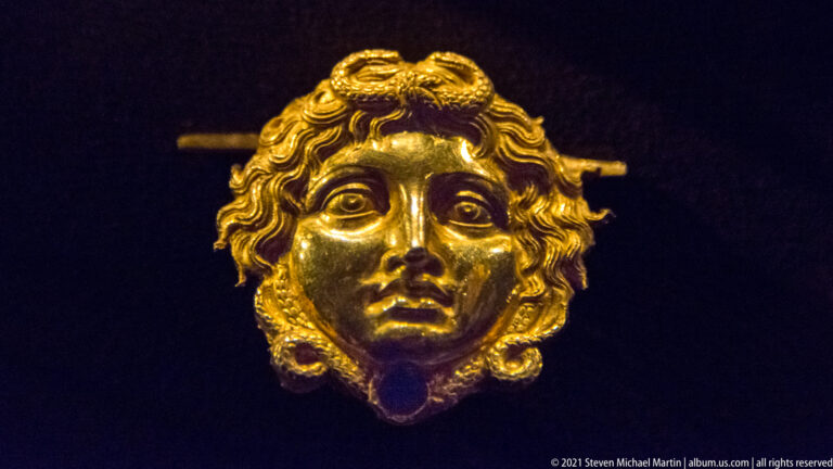 Small frontal gold face with large eyes, curling hair, and a horizontal attachment bar extending behind the head.