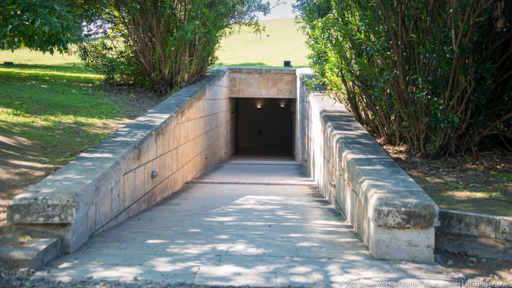 Paved ramp descending between stone retaining walls and greenery to a dark museum entrance beneath the landscape.