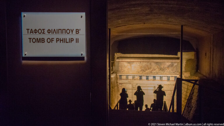Visitors descend through a dark museum passage toward the brightly illuminated façade of the Tomb of Philip II.