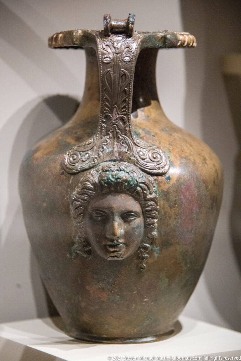Rounded bronze vessel with a relief female face below the handle and mottled green-brown patina across the body.