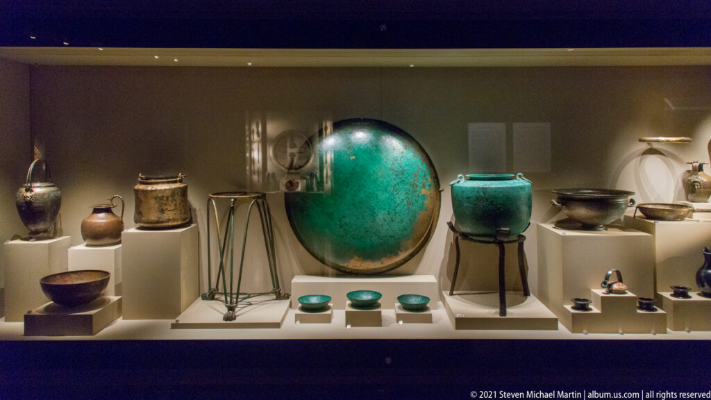 Museum case filled with metal bowls, jars, stands, and vessels around a large turquoise-patinated circular object.