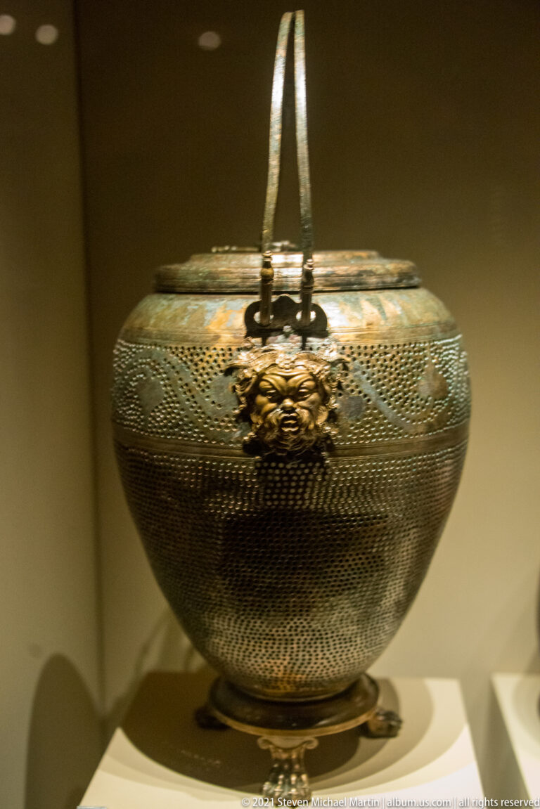 Rounded handled metal vessel with dense patterned surface, tall arched handle, front clasp, and patches of green corrosion.