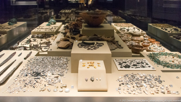Long museum case densely arranged with hundreds of small metal fragments, beads, ornaments, blades, and several larger vessels.