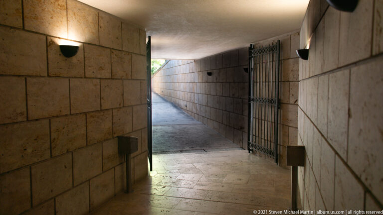 Narrow stone-clad museum corridor with wall lights, tiled floor, metal gate, and daylight at the far end.