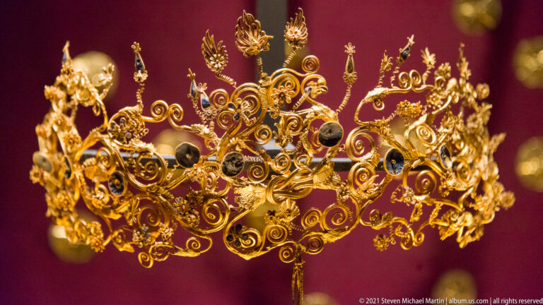 Elaborate gold wreath with curling wire tendrils, leaves, flowers, and dark spherical ornaments against a deep red background.