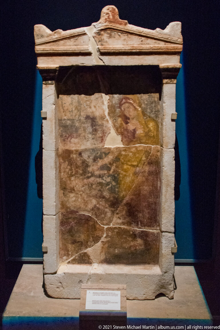 Small pedimented stone grave monument with a weathered multicolored panel containing fragmentary human figures.