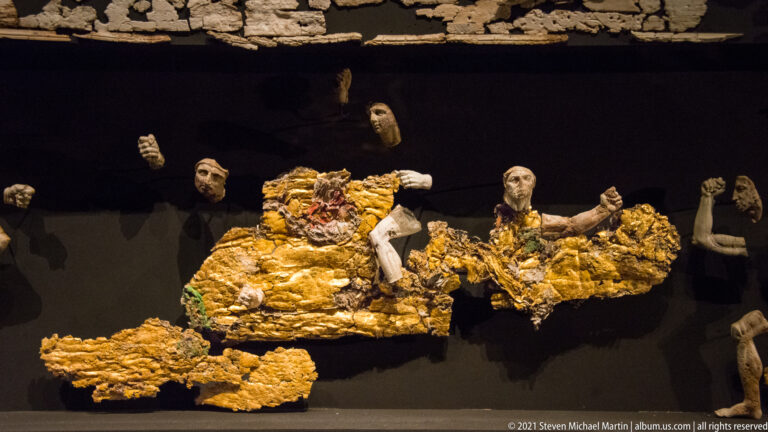 Fragmentary carved heads and limbs combined with broad gold-colored figure groups, mounted against a black museum background.