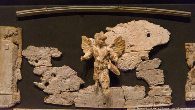 Small carved winged human figure mounted among broken pale plaques and fragments against a dark background.