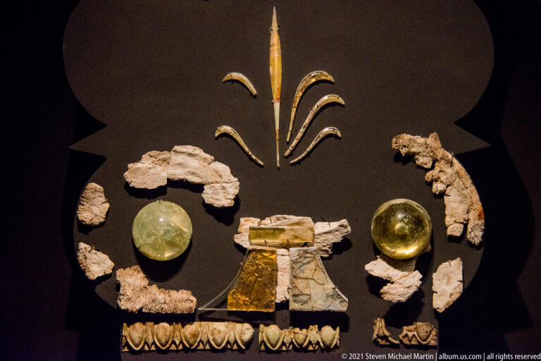 Assorted pale fragments, two translucent round disks, curved inlays, narrow pointed pieces, and gilded fittings arranged symmetrically on black.