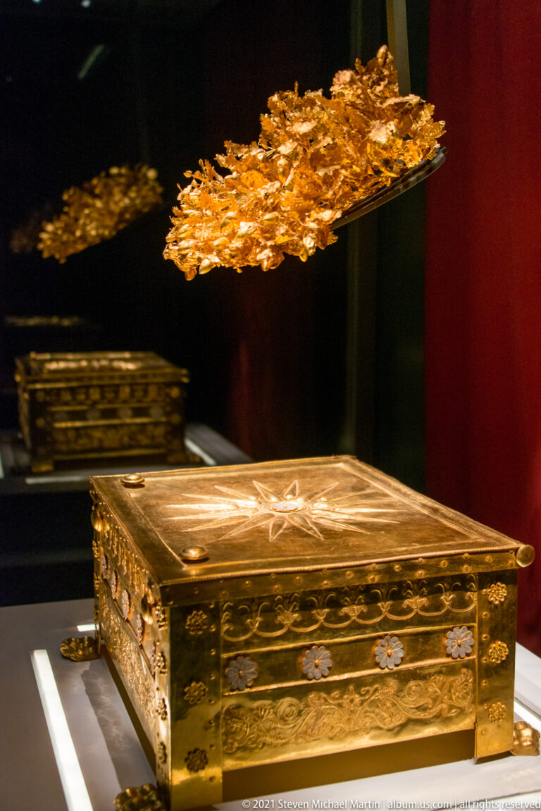 Gold larnax from the Tomb of Philip II displayed beneath its suspended gold oak wreath against a dark red background.