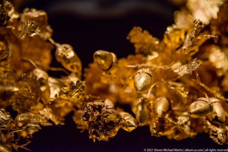 Macro view of a gold wreath with overlapping leaves, small buds, wire stems, and smooth oval fruit-like ornaments.