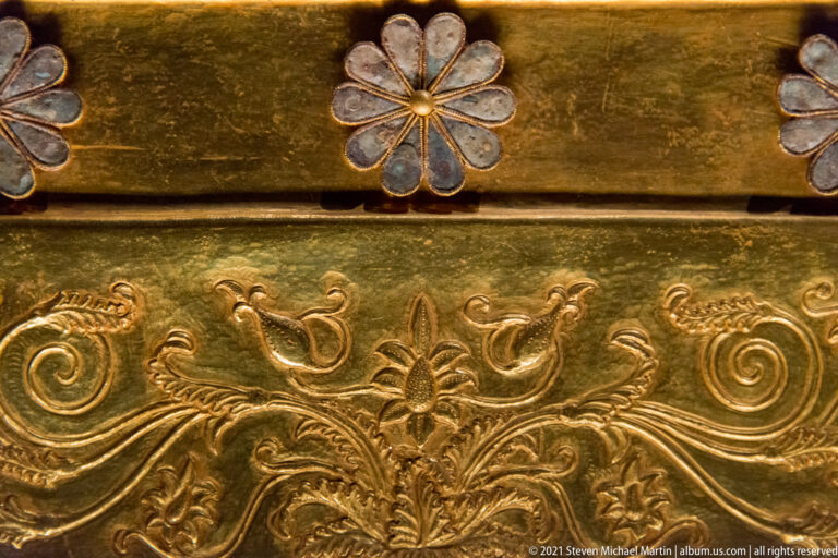 Close-up of gold chest ornament with scrolling foliage, animal forms, a central flower, and pale multi-petaled rosettes above.