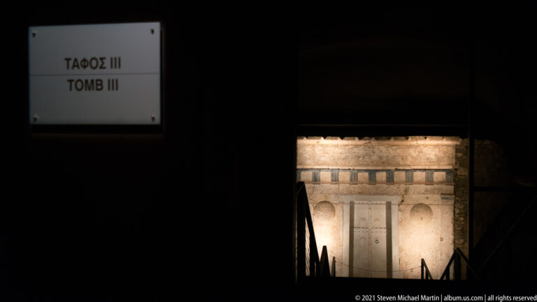Distant illuminated façade of Tomb III, the Prince’s Tomb, seen at the end of a dark underground museum gallery.