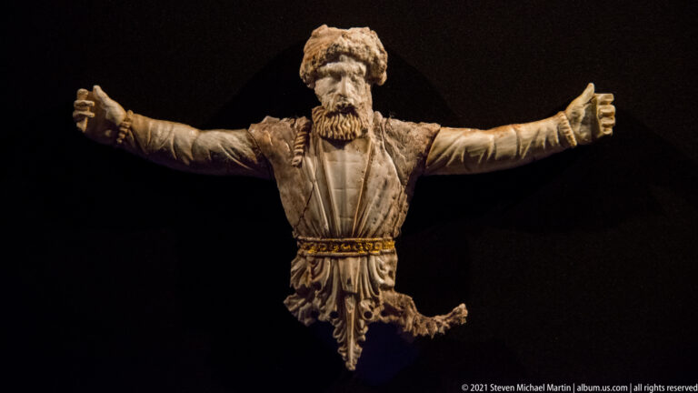 Carved bearded male figure with both arms stretched outward, belted drapery, and missing lower portions against black.